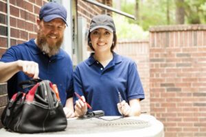 male-and-female-HVAC-technicians-wearing-blue-shirts-and-caps-outside-with-bag-of-tools-on-AC