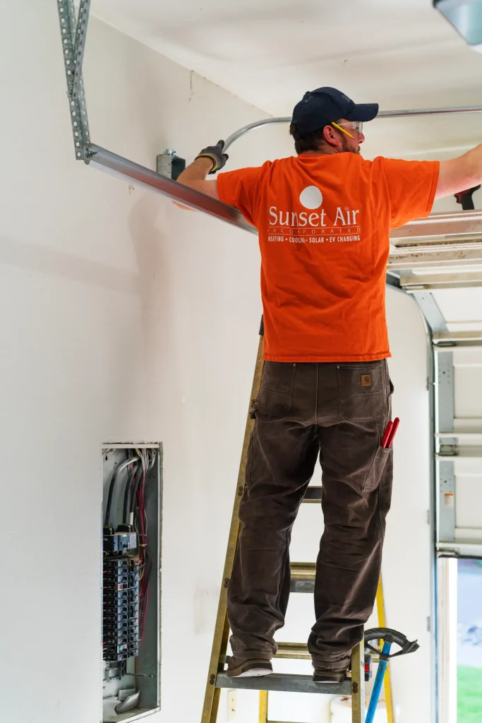 A technician in an orange shirt from Sunset Air is working on installing a metal track on a wall while standing on a ladder. The wall has an electrical panel and exposed wiring, indicating a home improvement or HVAC installation project. The technician is wearing safety glasses and gloves, emphasizing safety during the installation process. This image showcases professional HVAC services, highlighting the importance of skilled technicians in home maintenance and energy efficiency solutions.